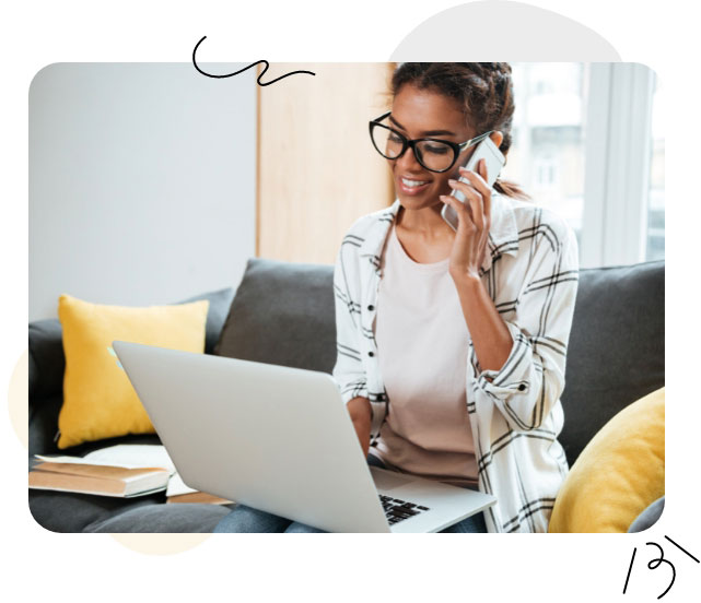 A woman in glasses sits on a couch, holding a laptop and a phone in her hands, multitasking efficiently.